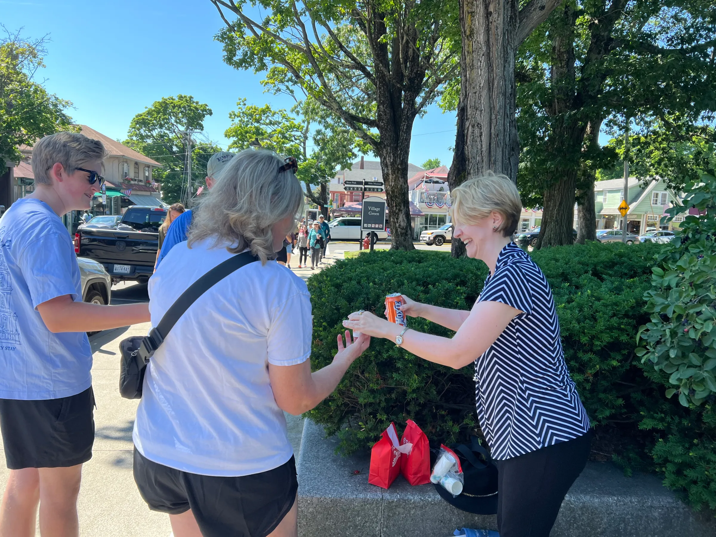 People exchanging items outdoors near trees and buildings on a sunny day.