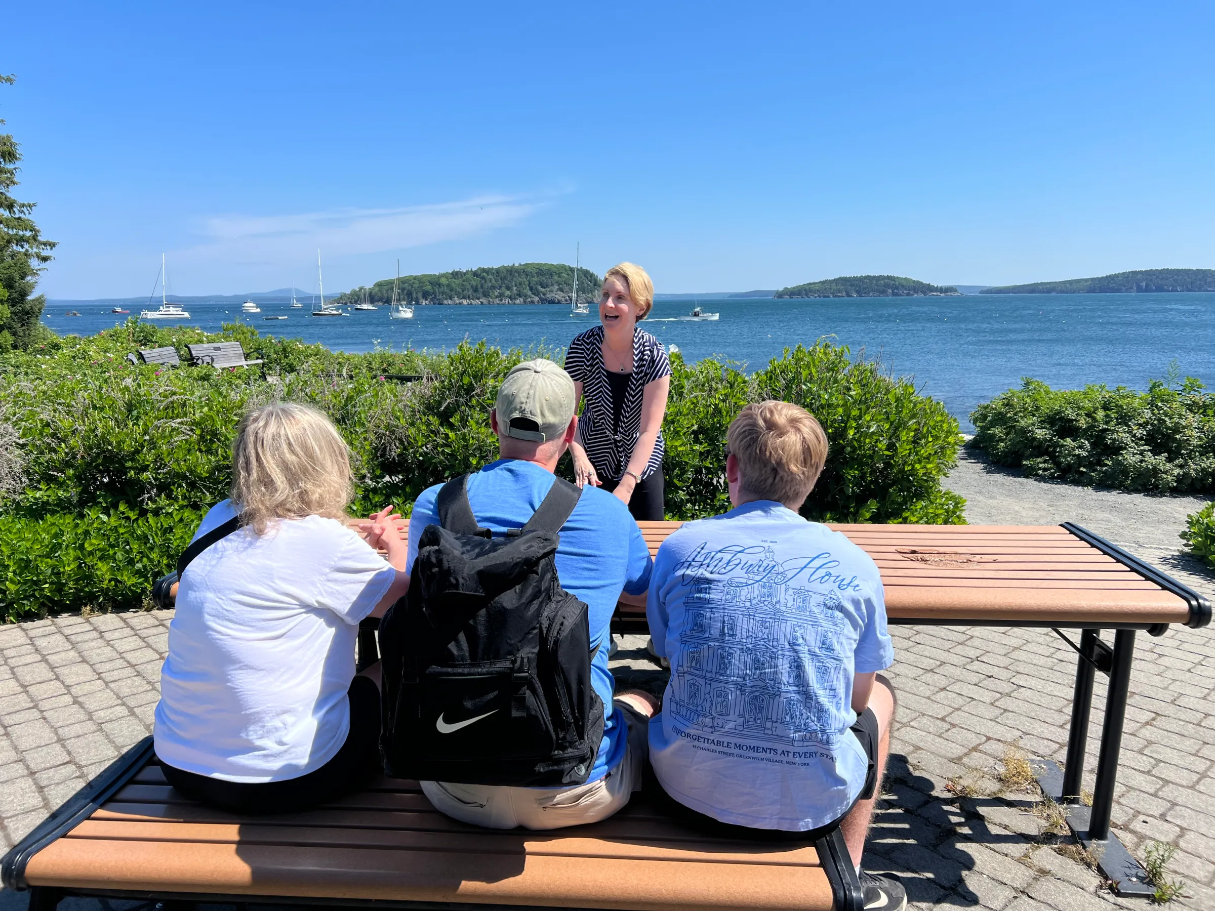 People sitting on bench facing woman with ocean and sailboats in background.