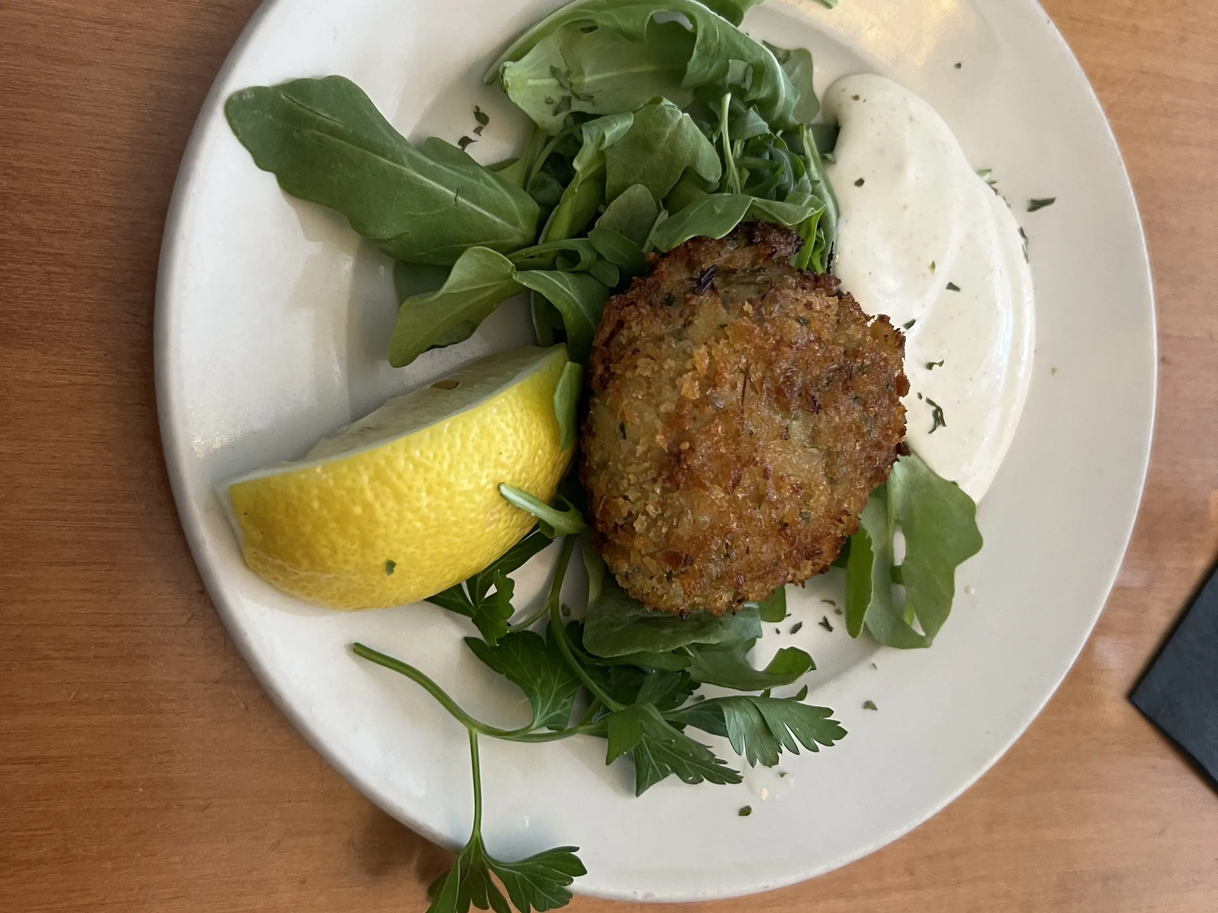 Fried crab cake on greens with lemon wedge and white sauce on a plate.