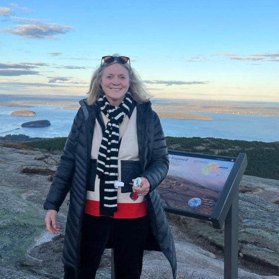 Woman in jacket and scarf stands on a scenic hilltop near an informational sign.