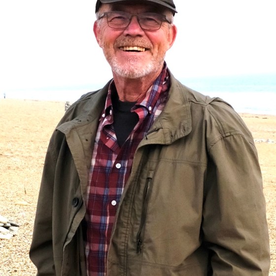 Smiling man wearing glasses and cap, standing on a beach.