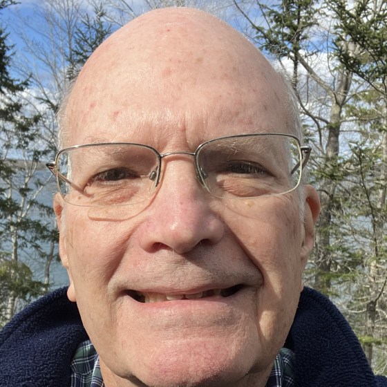 Smiling man with glasses outside, trees and sky in background.