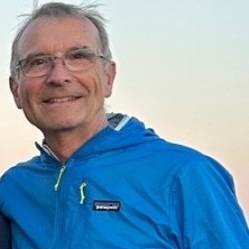 Smiling older man in blue jacket and glasses against a clear sky.