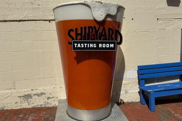 Large beer glass statue labeled 'Shipyard Tasting Room' on a pedestal, next to a blue bench.