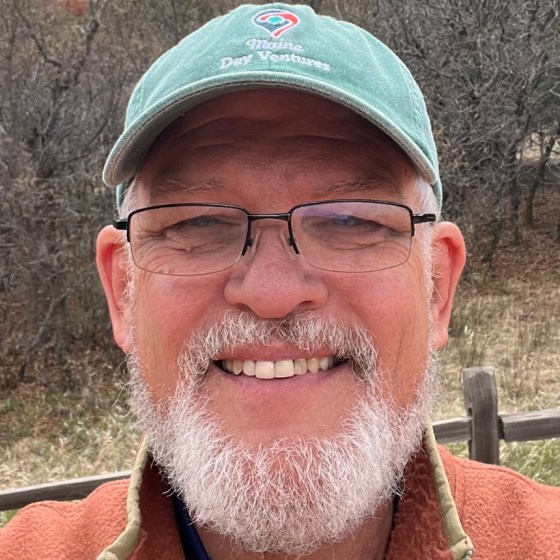 Smiling man with beard, glasses, and cap standing outdoors.