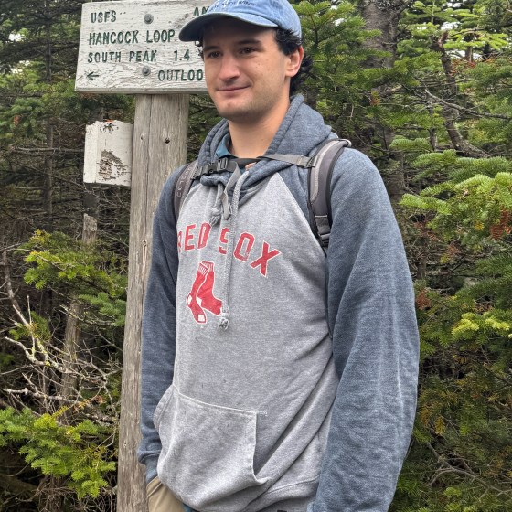 Person in a grey Red Sox hoodie stands near a forest trail sign.