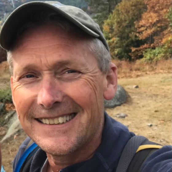Smiling person in outdoor setting with trees and hills in background.