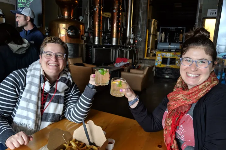 Two people smiling and toasting with drinks in a distillery.