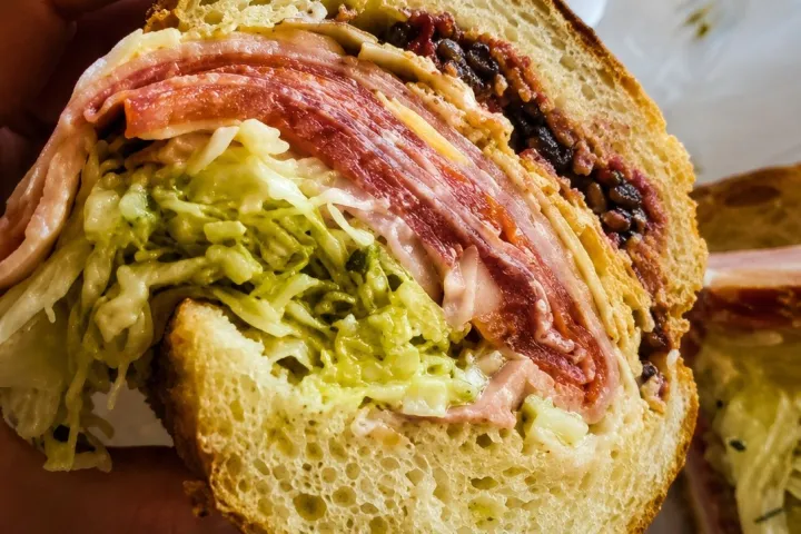 Hand holding a sandwich with meats, lettuce, and sauce.